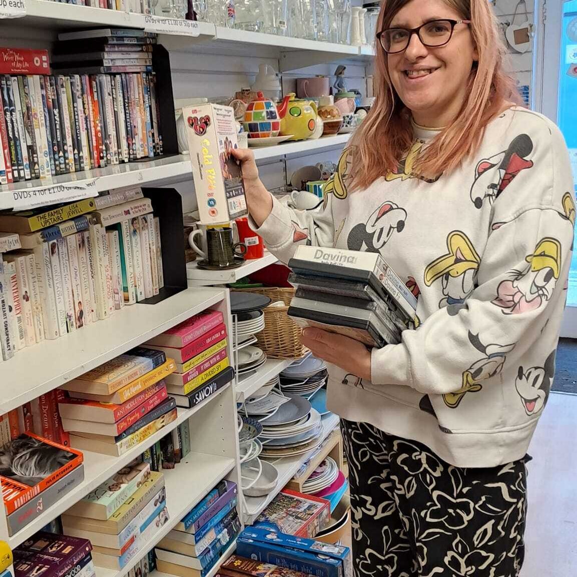 A lady smiling and sorting books on a shelf at the Style Acre Charity Shop