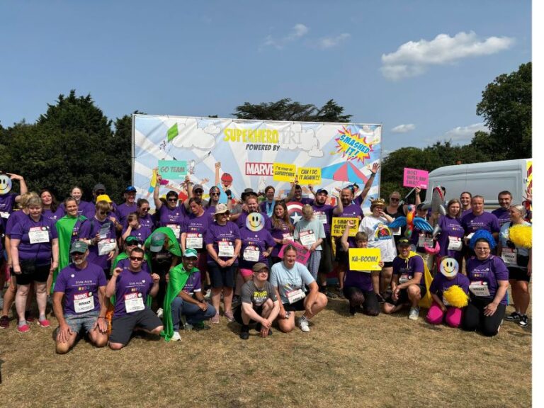 Superhero group (Liam, Donna and Kate blocked out) Group of 40 people standing, kneeling in front of a large Marvel Superhero sign celebrating their achievements at the Superhero Triathlon.