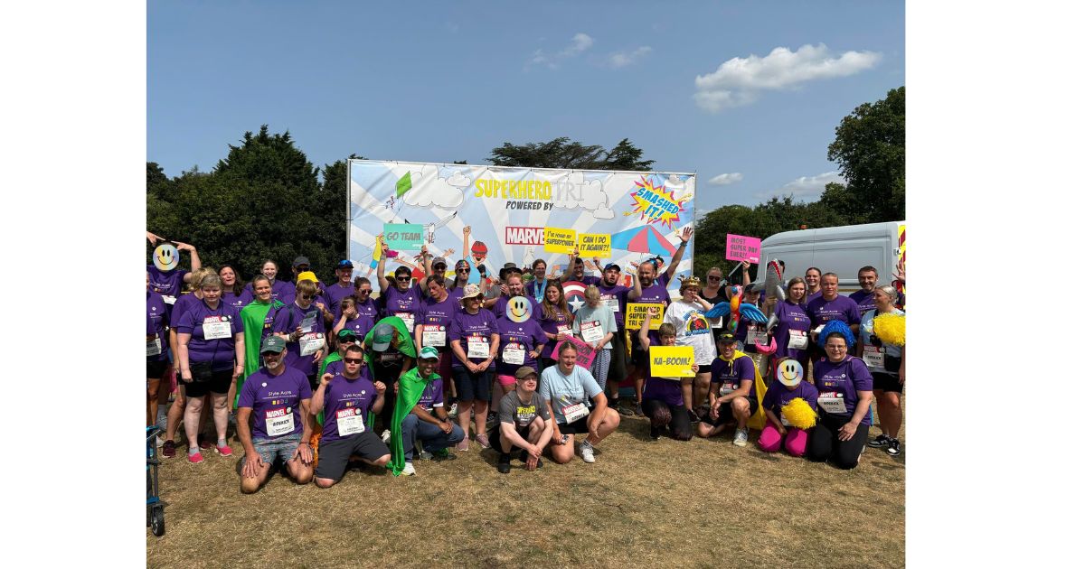 Superhero group (Liam, Donna and Kate blocked out) Group of 40 people standing, kneeling in front of a large Marvel Superhero sign celebrating their achievements at the Superhero Triathlon.
