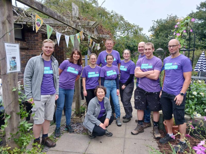 Group of 10 volunteers, men and women, from a local company standing in a garden wearing purple t-shirts.