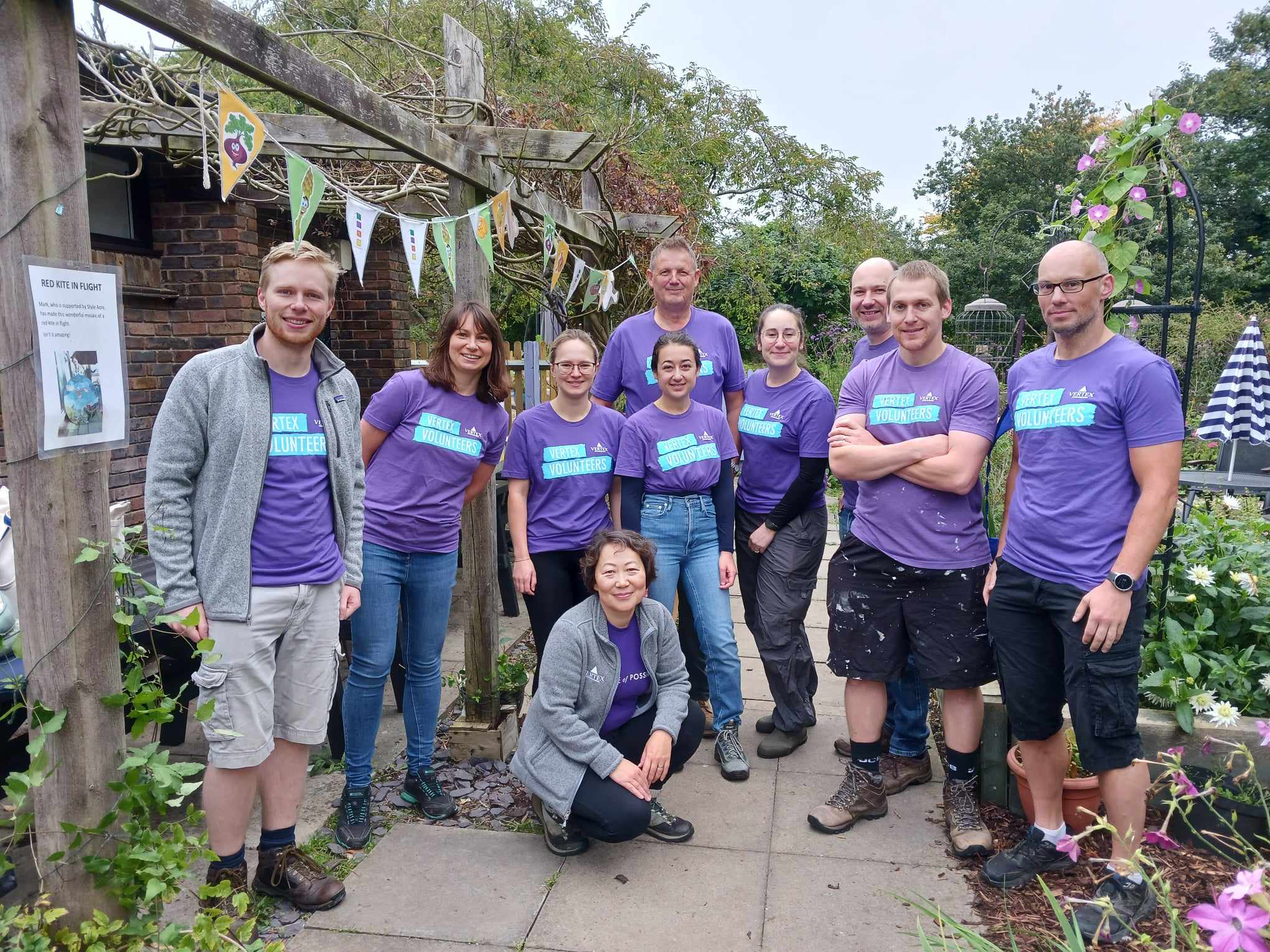 459247678_424950460175140_2361948515227169888_n Group of 10 volunteers, men and women, from a local company standing in a garden wearing purple t-shirts.