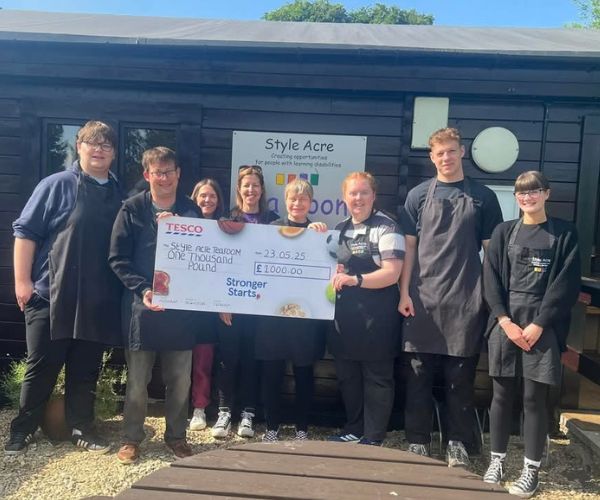 8 people standing outside the tea room holding a large cheque for £1000