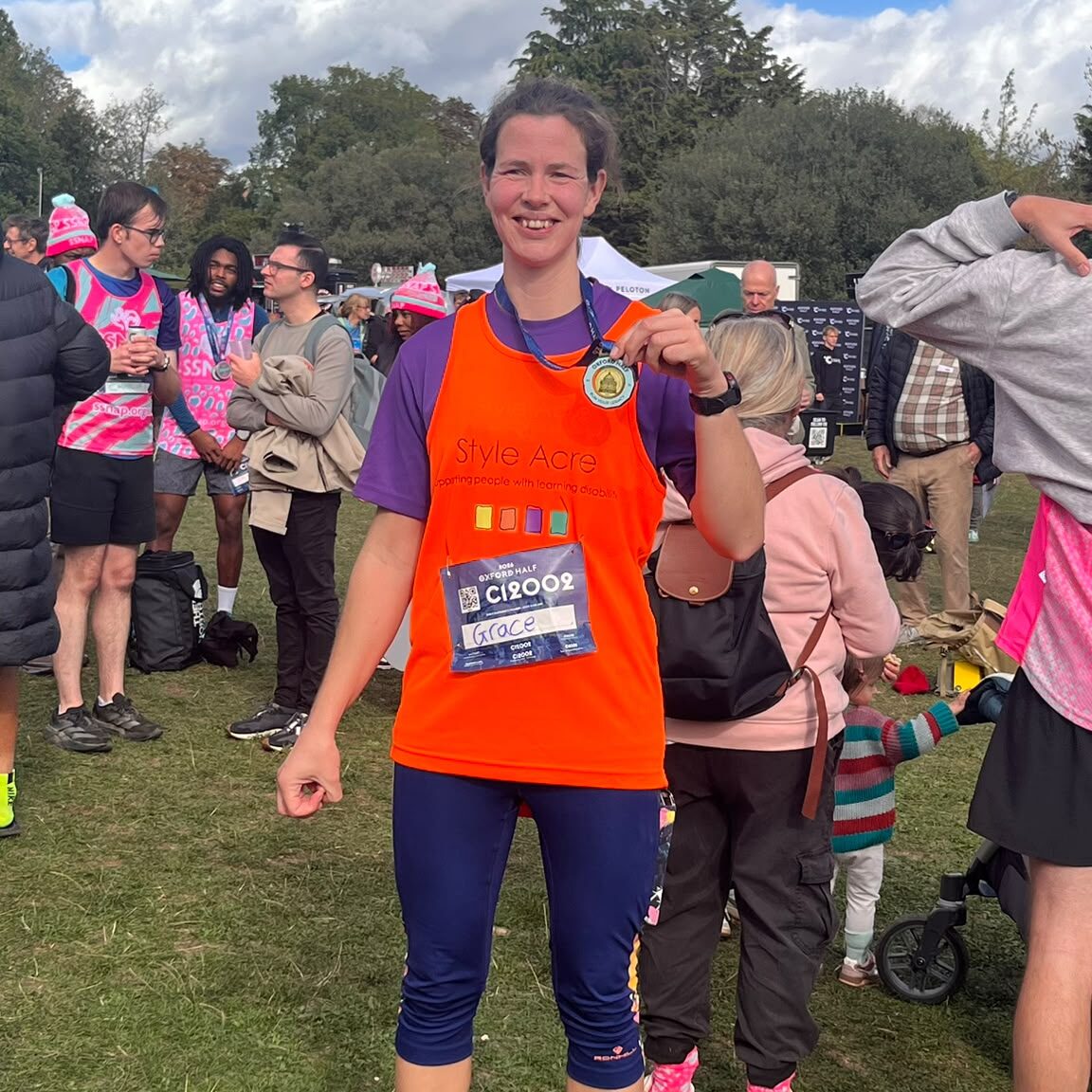 A lady standing in an orange running vest showing her Oxford Half Marathon medal