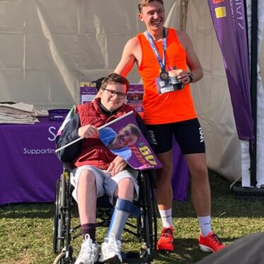 A man in a wheel chair holiday a cheering flag and a running in an orange vest holding his finish medal
